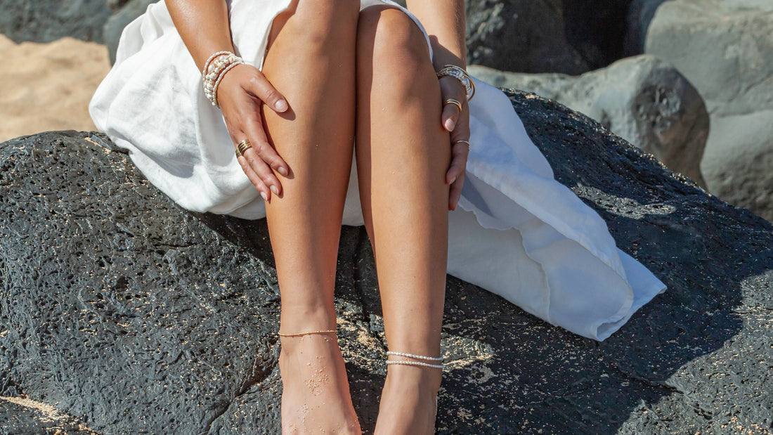 Person sitting on a rock by the beach with a white dress and sandals.